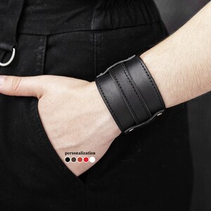 May include: A black leather wrist cuff with three horizontal straps and silver-tone rivets. The cuff is worn on a wrist, with a hand in a pocket. The word "personalization" is visible with color options.