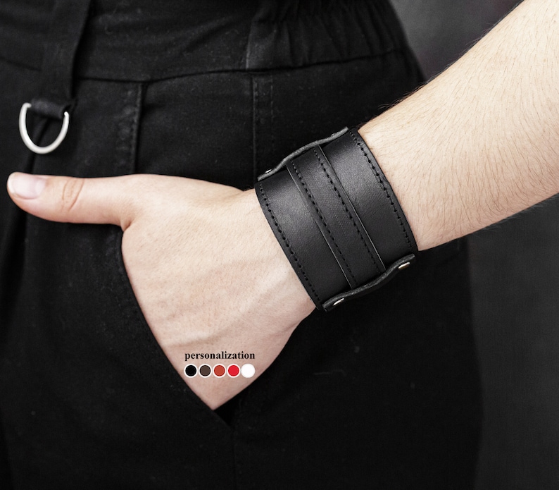 May include: A black leather wrist cuff with three horizontal straps and silver-tone rivets. The cuff is worn on a wrist, with a hand in a pocket. The word "personalization" is visible with color options.