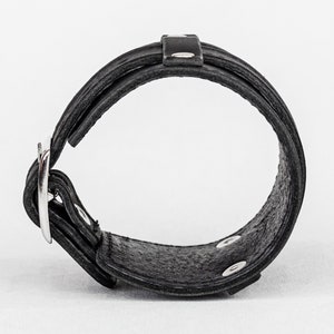 May include: Black leather bracelet with a silver buckle and snap closures. The bracelet features a layered design with a textured surface. The buckle is a simple, polished metal. The bracelet is designed to be worn around the wrist.