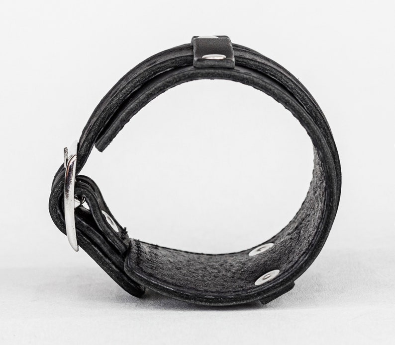 May include: Black leather bracelet with a silver buckle and snap closures. The bracelet features a layered design with a textured surface. The buckle is a simple, polished metal. The bracelet is designed to be worn around the wrist.