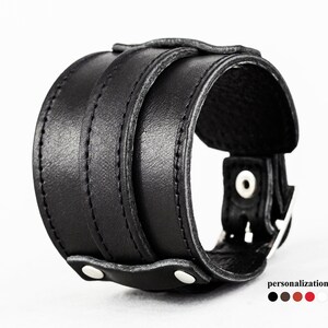 May include: A wide black leather cuff bracelet with a buckle closure and two rows of stitching. The bracelet has a sturdy, masculine design. The image also shows color options for personalization.