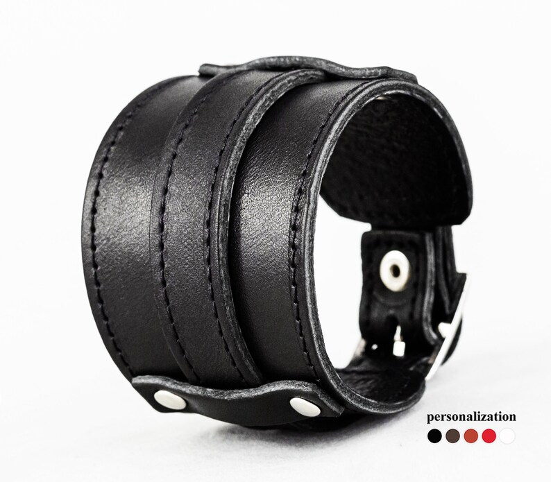 May include: A wide black leather cuff bracelet with a buckle closure and two rows of stitching. The bracelet has a sturdy, masculine design. The image also shows color options for personalization.