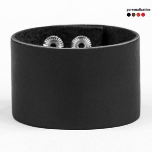 May include: A wide, black leather cuff bracelet with two silver-colored rivets. The bracelet is cylindrical and has a smooth texture. The word "personalization" is above a color palette of black, brown, red, and white.