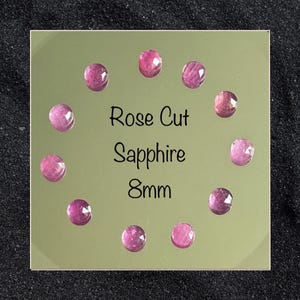 Pode incluir: Uma coleção de safiras lapidadas em rosa, cada uma com aproximadamente 8 mm de diâmetro, dispostas sobre uma superfície refletora. As pedras preciosas são de cor rosa profundo, com facetas que capturam a luz. O texto "Rose Cut Sapphire 8mm" é exibido.