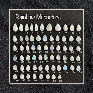 Puede incluir: Una exhibición de 63 piedras preciosas de Rainbow Moonstone en forma de lágrima, cada una numerada. Las piedras varían en tamaño y muestran una gama de colores, incluyendo blanco, azul y destellos iridiscentes. El texto "Rainbow Moonstone" está en la parte superior.