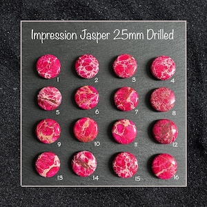 May include: A collection of sixteen drilled Impression Jasper gemstones, each approximately 1 inch in diameter, arranged on a dark surface. The stones are primarily a vibrant pink with intricate patterns of white and brown.