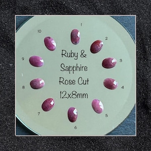 Peut inclure: Dix pierres précieuses rubis et saphir ovales, taille rose, disposées en cercle sur une surface claire. Chaque pierre est numérotée de 1 à 10. Le texte au centre indique Ruby & Sapphire Rose Cut 12x8mm. Les pierres sont rose foncé.