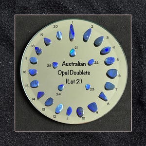 Puede incluir: Una pantalla circular de 25 dobletes de ópalo australiano, numerados del 1 al 25, sobre una superficie espejada. El texto "Australian Opal Doublets (Lot 2)" está impreso en el centro de la pantalla.