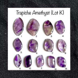 May include: A collection of thirteen Trapiche Amethyst gemstones, each with unique purple and white patterns. The gemstones are oval-shaped and numbered 1 through 13. The text "Trapiche Amethyst (Lot K)" is displayed at the top of the image.