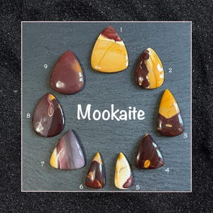 May include: Nine polished Mookaite stones in various shades of brown, red, and yellow, arranged in a circle on a black slate surface. The stones are labelled with numbers 1 through 9.