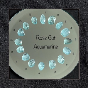 May include: Sixteen rose-cut aquamarine gemstones are displayed on a reflective surface, each numbered. The text "Rose Cut Aquamarine" is below the gemstones. The gemstones are a pale blue hue.