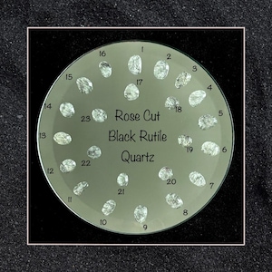 May include: A close-up of 23 rose cut black rutile quartz stones arranged in a circle on a white background. The stones are numbered 1 through 23.