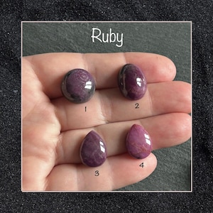 May include: Four polished ruby gemstones displayed on a palm. Two are oval-shaped, labeled 1 and 2, and two are teardrop-shaped, labeled 3 and 4. The stones exhibit deep magenta and purple hues with natural inclusions and reflective surfaces.