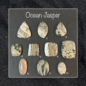 Może przedstawiać: Kolekcja dziesięciu kamieni Ocean Jasper w różnych kształtach, w tym łezki, owalne i prostokątne. Kamienie prezentują mieszankę odcieni beżu, brązu i zieleni z unikalnymi wzorami. Tekst "Ocean Jasper" jest widoczny na górze.