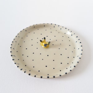 May include: A small, round ceramic plate with a cream-colored base and scattered navy blue polka dots. The rim is dotted with navy blue paint. A small, yellow ceramic animal figure is in the center of the plate.