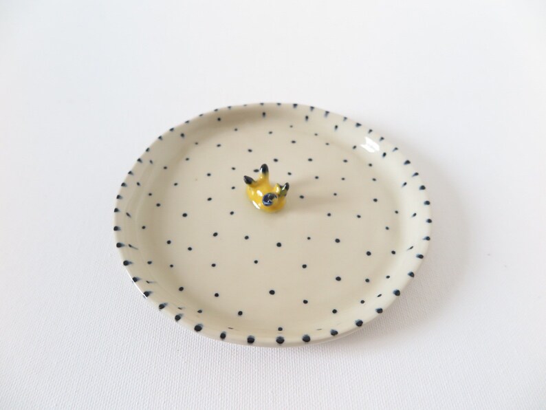 May include: A small, round ceramic plate with a cream-colored base and scattered navy blue polka dots. The rim is dotted with navy blue paint. A small, yellow ceramic animal figure is in the center of the plate.