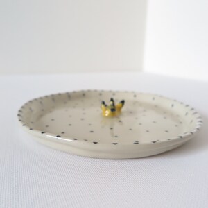 May include: A small, off-white ceramic dish with a dotted pattern and a decorative yellow and black floral accent. The rim is detailed with a blue dotted border. The dish is oval-shaped.