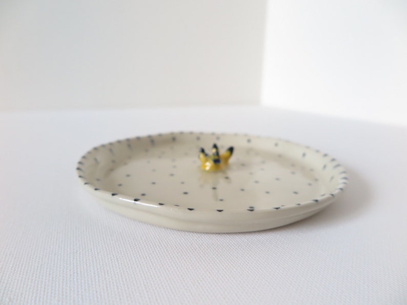 May include: A small, off-white ceramic dish with a dotted pattern and a decorative yellow and black floral accent. The rim is detailed with a blue dotted border. The dish is oval-shaped.