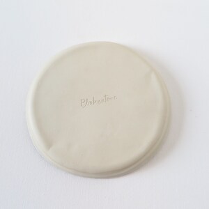 May include: A round, off-white ceramic coaster with a slightly uneven surface. The word "Pukahutara" is subtly inscribed on the coaster. The coaster is designed to protect surfaces from drink condensation.