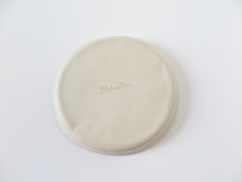 May include: A round, off-white ceramic coaster with a slightly uneven surface. The word "Pukahutara" is subtly inscribed on the coaster. The coaster is designed to protect surfaces from drink condensation.