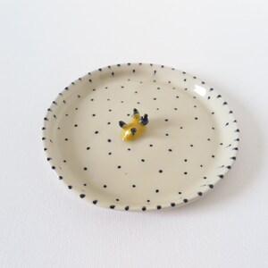 May include: A small, round, off-white ceramic plate with a pattern of small black dots. The rim is also decorated with black dashes. A small, yellow, abstract sculpture with black accents sits in the center of the plate.