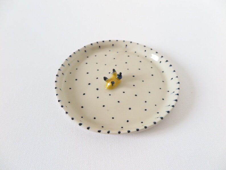 May include: A small, round, off-white ceramic plate with a pattern of small black dots. The rim is also decorated with black dashes. A small, yellow, abstract sculpture with black accents sits in the center of the plate.