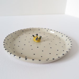 May include: A small, off-white ceramic dish with a raised yellow and black decorative element in the center. The dish is dotted with small, evenly spaced black spots and has a scalloped edge.