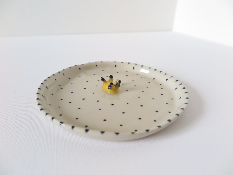 May include: A small, off-white ceramic dish with a raised yellow and black decorative element in the center. The dish is dotted with small, evenly spaced black spots and has a scalloped edge.