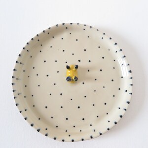 May include: A cream-colored ceramic plate with a yellow and blue animal figurine in the center. The plate is decorated with small, evenly spaced black dots and a border of dark blue dashes.
