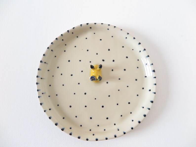 May include: A cream-colored ceramic plate with a yellow and blue animal figurine in the center. The plate is decorated with small, evenly spaced black dots and a border of dark blue dashes.