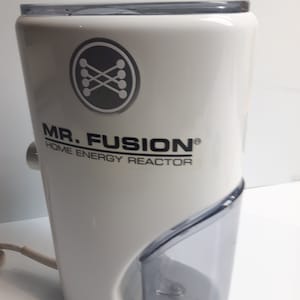 Pode incluir: Um "Mr. Fusion Home Energy Reactor" branco com um compartimento de plástico transparente e um logótipo cinzento. O dispositivo tem um topo arredondado e um cabo de alimentação. O logótipo apresenta um design estilizado de linhas e círculos que se cruzam.