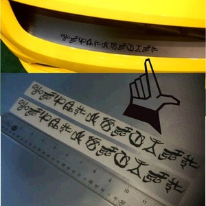 May include: A yellow car bumper with a black decal featuring stylized Japanese characters. Two additional decals and a ruler are shown below. A black hand graphic with an extended index finger is also visible.