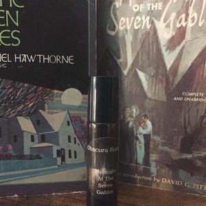 May include: A black roll-on bottle of perfume with a white label that reads "Obscura Horti Midnight At The Seven Gables". The bottle is sitting on a wooden surface in front of two books. The book on the left has the title "The House of the Seven Gables" and the book on the right has the title "The House of the Seven Gables".