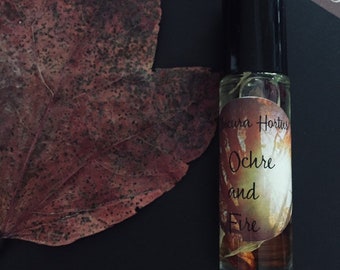 May include: A small glass bottle with a black cap and a label that reads "Ocura Hortus Ochre and Fire". The bottle is filled with a clear liquid and contains dried flowers and herbs.