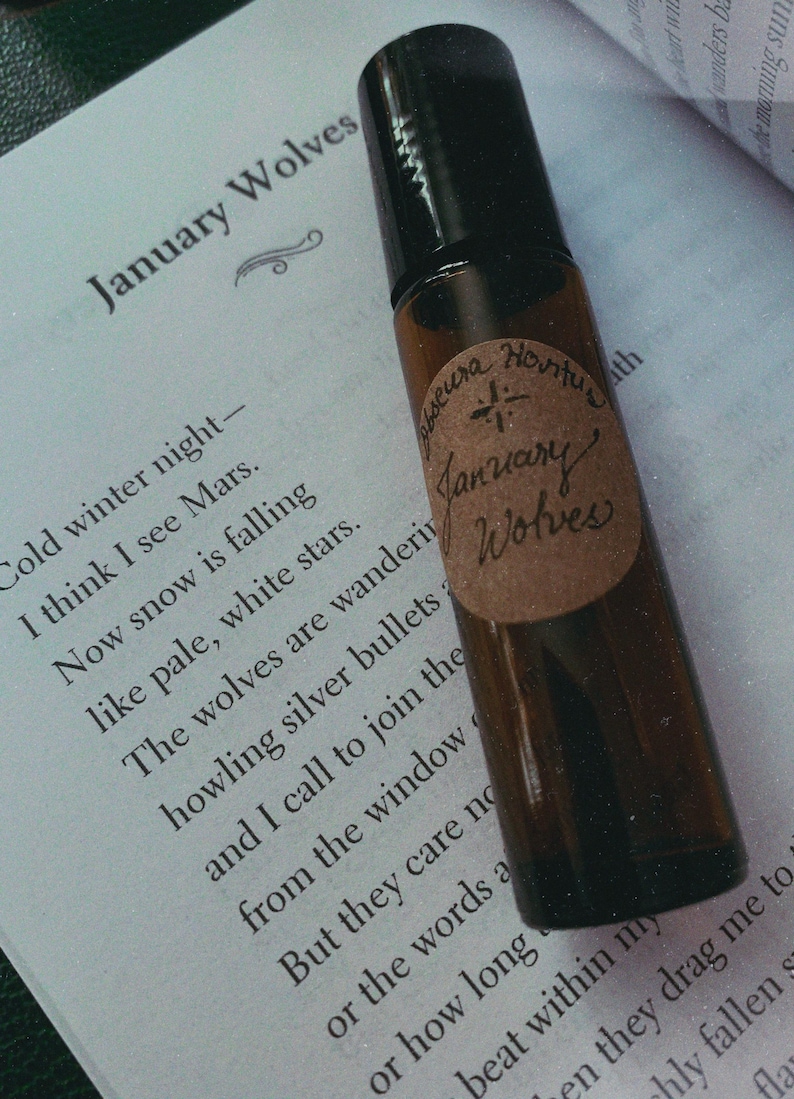 May include: A brown glass essential oil roller bottle with a black cap, labeled "January Wolves" in handwritten script. The bottle rests on a page of a book with a poem about winter. The label also includes the words "Obseura Hortus".