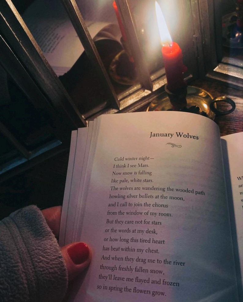 May include: An open book with the title "January Wolves" is held near a lit red candle. The poem's text is visible, and the scene is dimly lit, creating a cozy, intimate atmosphere. A mirror reflects the candle's flame.