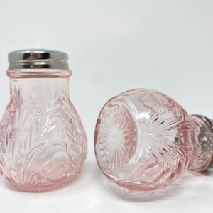 May include: Two pink glass salt and pepper shakers with silver tops. The shakers have a floral design and are in focus. The background is white.