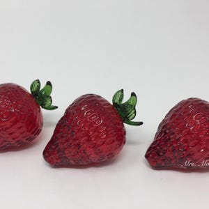 May include: Three red glass strawberries with green glass leaves. The strawberries are textured to look like real strawberries.