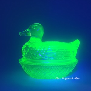 May include: A green glass duck shaped dish with a woven basket design. The dish is sitting on a nest shaped base.
