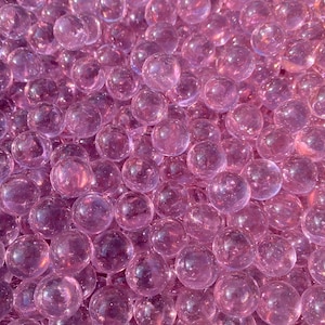 May include: A close-up shot of numerous transparent, pink glass marbles. The marbles are clustered together, creating a dense, textured surface. The image is well-lit, highlighting the marbles' round shape and glossy finish. The text "Mrs. Margaret's Glass" is visible.
