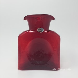 May include: A vibrant red glass vase with a square base and a rounded, flared top. The vase has a smooth, glossy finish and a label that reads "Blenco Handcrafts Made in USA". The overall shape is reminiscent of a flask.