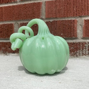 May include: A light green glass pumpkin with a curled stem. The pumpkin has a smooth, glossy finish and is sitting on a light gray surface.