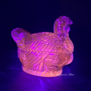May include: A clear glass turkey-shaped dish with a lid. The dish is a pale pink color under a black light.