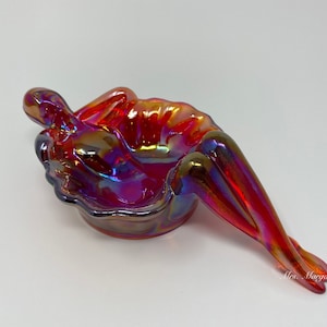 May include: Iridescent red glass figurine in the shape of a reclining figure. The glass has a glossy finish with hues of red, purple, and gold. The figurine is a decorative art piece.