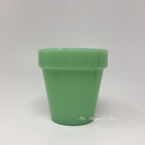 May include: A light green glass flower pot with a slightly flared shape. The pot has a smooth, glossy finish and a small, raised rim. The text "Mrs. Margaret's Glass" is visible on the lower right side.