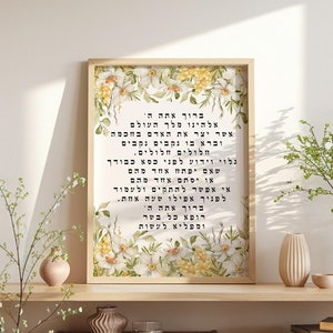 May include: A framed print with text in Hebrew, surrounded by a floral border of yellow and white flowers. The print is displayed on a wooden cabinet with decorative items and greenery.