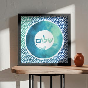 May include: A framed print featuring a blue and green watercolour circle design. The circle contains the Hebrew word "Shalom" written in white. The background is a pattern of blue dots.