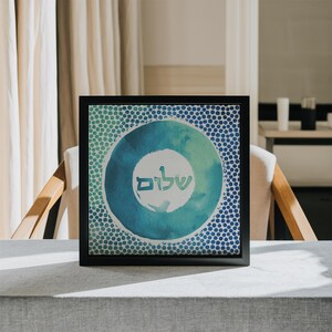 May include: A framed print featuring a blue and green watercolour circle design. The circle has a white background with the Hebrew word "Shalom" written in black.