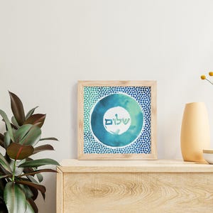 May include: A framed print featuring a blue and green watercolour circle design. The circle contains the Hebrew word "Shalom" written in white. The frame is made of wood and has a light brown finish.
