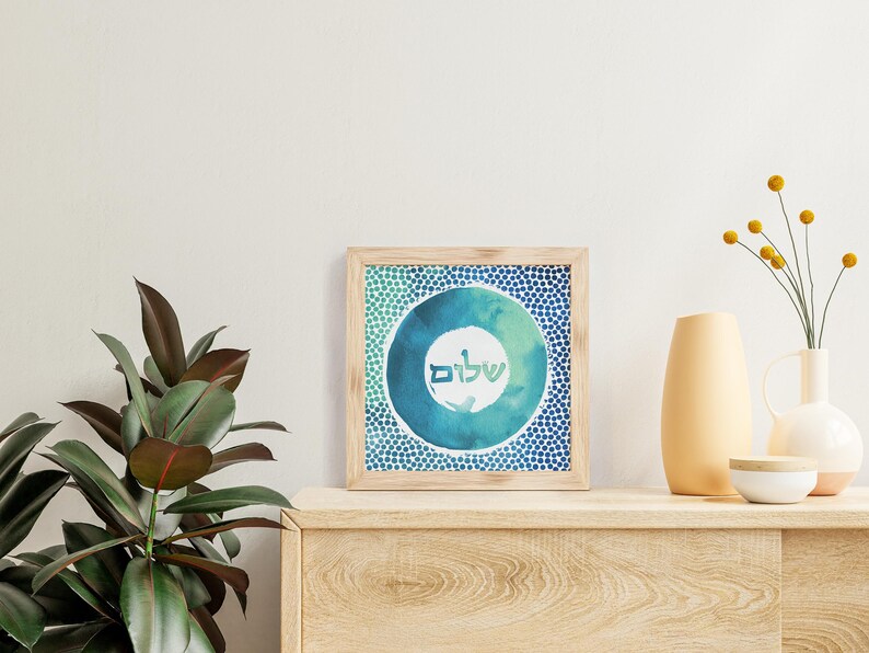May include: A framed print featuring a blue and green watercolour circle design. The circle contains the Hebrew word "Shalom" written in white. The frame is made of wood and has a light brown finish.